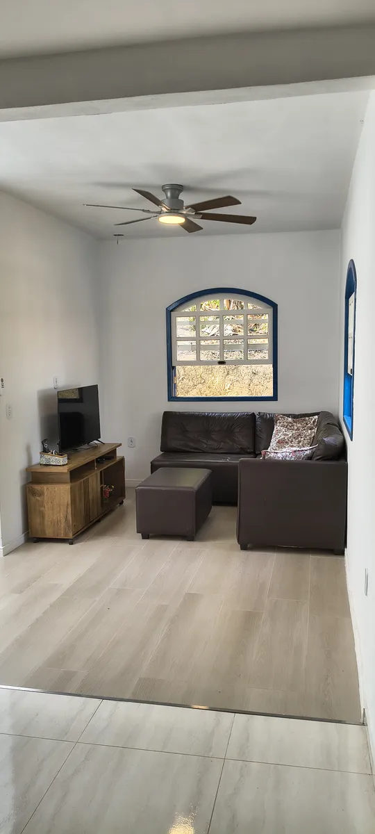 Cantinho aconchegante da sala com sofá de canto, TV e ventilador de teto com janela colonial no Sítio Canto da Seriema